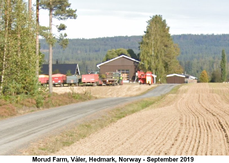 The Morud Farm, Våler, Hedmark, Norway: Color photo taken in September 2019 of farm outbuildings standing behind a row of modern farm wagons along a gravel road that runs along a freshly plowed field, with a wooded hillside in the distance